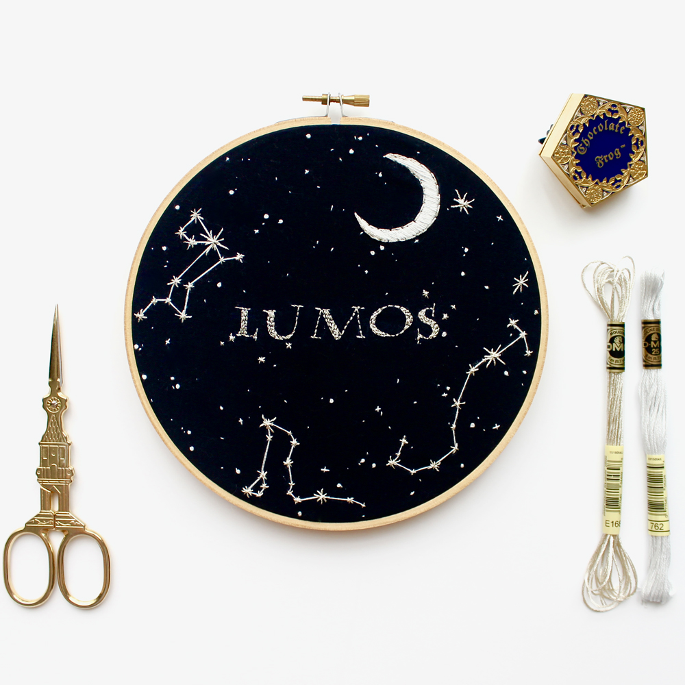 flat lay of modern embroidery hoop with harry potter design featuring lumos spell, chocolate frog pin, scissors and metallic embroidery thread